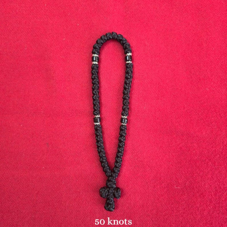 May include: A black knotted cord prayer rope featuring fifty individual knots arranged in a continuous loop. The rope includes small silver-colored spacer beads positioned at intervals and terminates in a knotted cross at the bottom.