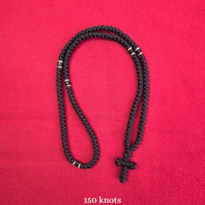 May include: A black knotted cord necklace featuring a cross pendant at the base. The cord is constructed from 150 knots and includes small silver-toned metallic accents spaced along the length of the circular loop, creating a textured design.