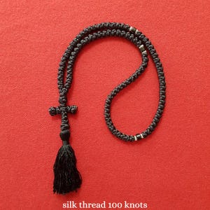 May include: A black knotted prayer rope necklace featuring a cross-shaped knot, a long tassel, and small metallic beads integrated into the cord. The necklace is composed of silk thread and contains 100 knots as indicated by the text at the bottom.