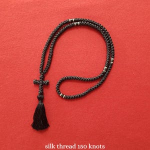 May include: A black knotted prayer rope necklace featuring a cross pendant and a tassel at the bottom. The cord is constructed from black thread with 150 knots and includes small metallic silver-colored beads integrated into the circular design.