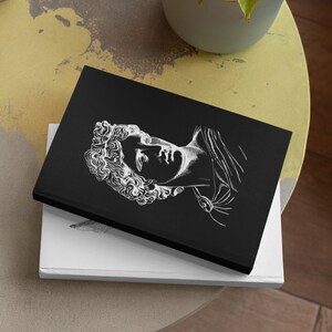 May include: A black notebook with a white line drawing of a classical sculpture of a person's head and shoulders. The notebook is sitting on a white surface.