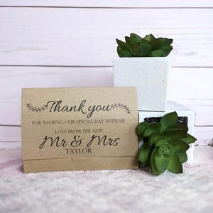 Könnte beinhalten: Eine Dankeskarte aus Kraftpapier mit dem Text "Thank you" und "Mr & Mrs Taylor". Die Karte ist mit floralen Akzenten verziert. Die Karte wird mit weißen quadratischen Pflanzgefäßen und grünen Sukkulenten präsentiert.