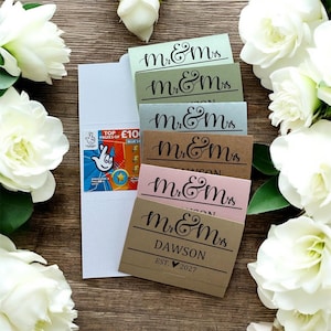 Könnte beinhalten: Eine Auswahl an Hochzeitskarten in Pastellfarben, jede mit dem Schriftzug "Mr & Mrs" in Schreibschrift. Eine Karte ist mit "Dawson" und "EST. 2027" personalisiert. Ein weißer Umschlag enthält ein Lotterielos. Weiße Blumen umgeben die Karten.