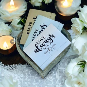 Op de afbeelding: Een houten hartvormige kom gevuld met drie kleine kaarten. De kaarten hebben de woorden "LOVE" en "LOVE always wins" in zwart schrift. De onderste kaart heeft de namen "Gavin & Stacey" en de datum "07-10-25". Witte rozen en brandende kaarsen omringen de kom.