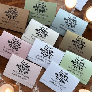 Könnte beinhalten: Eine Ansammlung von Streichholzschachteln in verschiedenen Pastellfarben, jeweils mit dem Aufdruck "lucky in love", den Namen "MR & MRS WATKINS" und dem Datum "Established. 14.10.27" in schwarzer Schrift. Die Schachteln liegen auf einer Holzoberfläche.