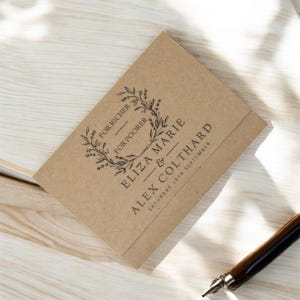 Op de afbeelding: Een gevouwen trouwuitnodiging van kraftpapier met zwarte tekst en een bloemenkrans. De tekst bevat "For Richer, For Poorer", de namen "Eliza Marie & Alex Colthard" en de datum "Saturday 19th September". Een pen is zichtbaar.