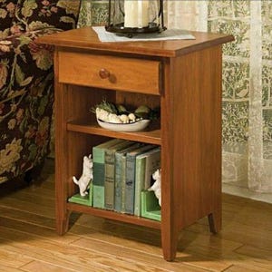 May include: A wooden end table with a drawer, two shelves, and a light wood finish. The table has a pair of white ceramic bookends shaped like dogs on the bottom shelf.