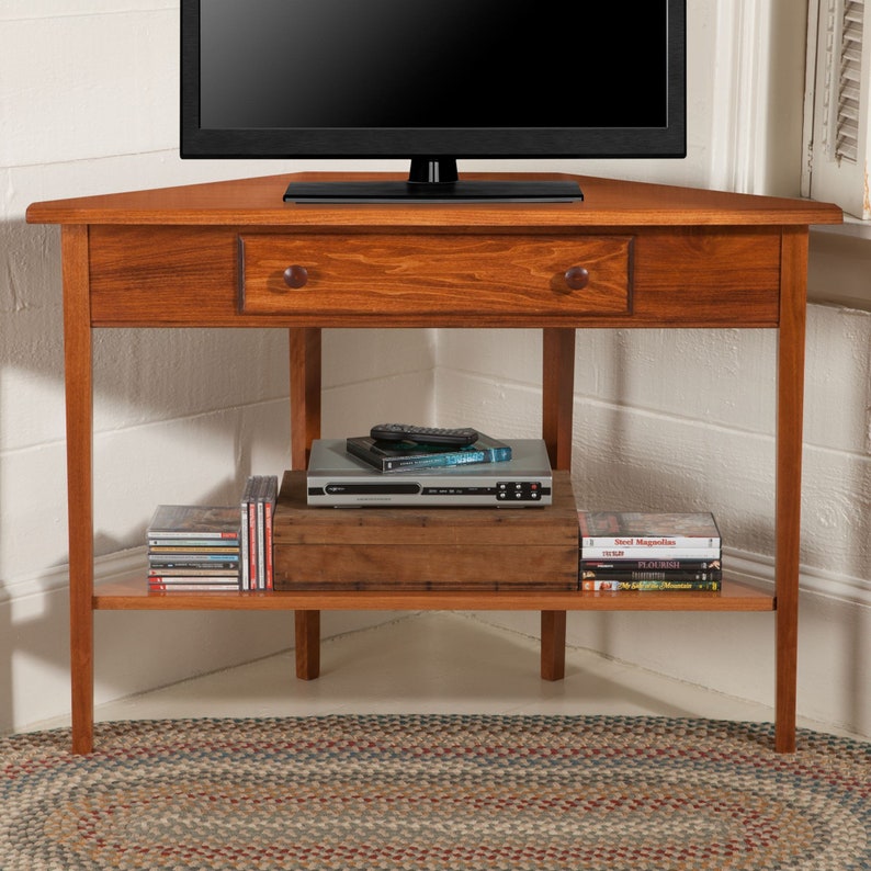 May include: A corner TV stand made of solid wood with a drawer and a shelf. The stand is stained a rich brown color and has a simple, traditional design. It is perfect for displaying a TV and other electronics.