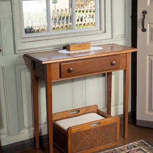 May include: A wooden side table with a drawer and a lower shelf. The table is a warm brown color and has a small wooden box on top. A woven basket with a cushion is placed on the lower shelf. The table is 30 inches tall.