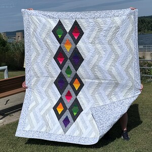 May include: A square quilt featuring a central vertical column of black diamond shapes filled with vibrant neon green, pink, orange, and purple fabric. The surrounding areas consist of white and gray chevron patterns with a light gray patterned border.