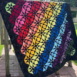 May include: A quilt featuring a vibrant rainbow gradient of fabric pieces arranged in a geometric star pattern against a solid black background. The design transitions through shades of red, orange, yellow, blue, and purple, creating a spectrum effect.