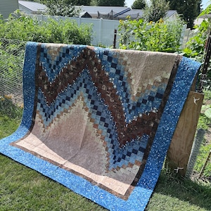 May include: A large rectangular quilt featuring a chevron pattern composed of small square fabric patches in shades of blue, brown, and beige. The quilt has a wide blue border with a mottled texture and visible quilted stitching throughout.