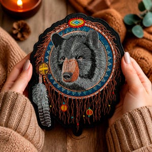 May include: A black embroidered patch featuring a detailed bear head centered within a circular frame. The design includes a blue beaded-style border, a top geometric emblem, and hanging brown fringe threads with a single grey feather accent.
