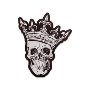 Pode incluir: Um patch preto e branco com uma caveira usando uma coroa. A caveira tem sombreamento detalhado e um sorriso largo. A coroa é ornamentada com flor-de-lis e detalhes de joias. O patch tem uma borda preta.