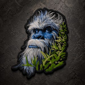 May include: Embroidered patch featuring a blue and white Sasquatch face with a long white beard and blue eyes. The patch is surrounded by green foliage and has a black border. The design is detailed and artistic.