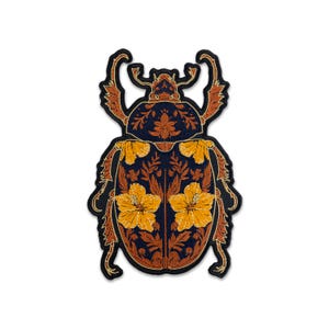 May include: An embroidered patch shaped like a beetle with a dark navy blue base. The insect features intricate orange and gold thread detailing, including floral patterns and two prominent yellow hibiscus flowers on its wing covers.