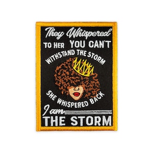 Peut inclure: Un écusson brodé rectangulaire avec un fond noir et une bordure jaune. Il présente le dessin d'une personne aux cheveux bruns bouclés portant une couronne jaune, avec le texte : They Whispered TO HER YOU CAN'T WITHSTAND THE STORM She whispered back I am THE STORM.