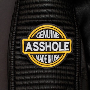 Puede incluir: Un parche bordado circular con fondo negro, bordes amarillos y texto blanco. El parche muestra la palabra GENUINE en un arco en la parte superior, ASSHOLE en una barra horizontal central y MADE IN USA en un arco en la parte inferior.