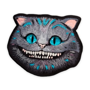 May include: Embroidered patch featuring the Cheshire Cat from Alice in Wonderland. The cat has a gray face with blue stripes, large blue eyes, a pink nose and ears, and a wide grin with white teeth. The patch has a black border.
