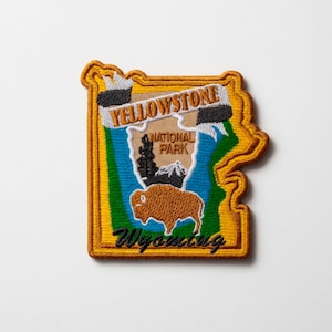 Op de afbeelding: Geborduurde patch met een kaartomtrek van Wyoming met een bizon, bergen en bomen. De patch leest "Yellowstone National Park" en "Wyoming" in een bannerontwerp, met een gouden rand.