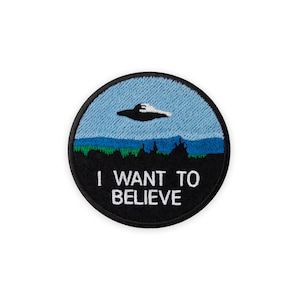 Könnte beinhalten: Runder, gestickter Aufnäher mit einem UFO über einer Landschaft und dem Text „I WANT TO BELIEVE“. Der Aufnäher hat einen schwarzen Rand, einen blauen Himmel und eine grüne und blaue Landschaft.