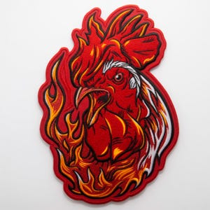 Pode incluir: Patch bordado com a imagem de uma cabeça de galo vermelho envolta em chamas. O galo tem um design detalhado com detalhes brancos e pretos, e as chamas são em tons de amarelo e laranja. O patch tem uma borda vermelha.