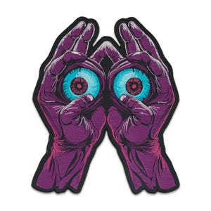 Puede incluir: Parche bordado con dos manos moradas formando una forma de binocular, cada mano sostiene un gran ojo azul con un iris rojo. El parche tiene un borde negro y una apariencia texturizada.