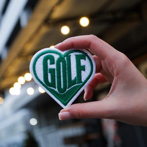 Puede incluir: Un parche en forma de corazón con la palabra "GOLF" bordada en verde sobre un fondo blanco. El parche se sostiene frente a un fondo borroso.