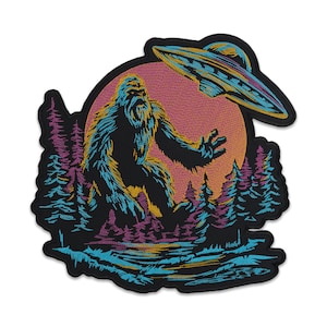 Op de afbeelding: Geborduurde patch met Bigfoot die naar een UFO reikt. Het ontwerp omvat een roze en oranje zonsondergang, bomen en een meer. De patch, met een zwarte rand, is geschikt voor jassen, tassen of andere stoffen items.