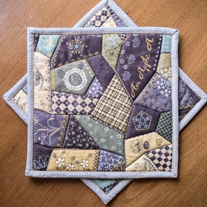 May include: A patchwork quilt hot pad with a blue and white border. The quilt is made of various fabrics in shades of purple, green, and brown. The text "An Apple A" is stitched in the center of the quilt.