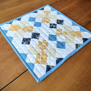 May include: A small blue and white quilted table runner with a diamond pattern and yellow floral fabric accents.