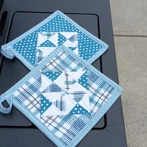 May include: Two blue and white quilted pot holders with a geometric pattern. The pot holders are on a black surface.