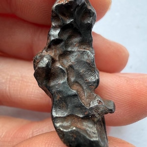 May include: A dark, irregularly shaped, metallic-looking object with a rough, textured surface. The object has a dark brown color with some areas appearing almost black. The overall shape is elongated and somewhat organic, with ridges and uneven edges.