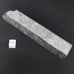 May include: A gray, rectangular meteorite specimen with a geometric Widmanstätten pattern. A small white cube with the text "1cm" is next to the meteorite. The meteorite has a rough, textured surface and is displayed on a dark background.