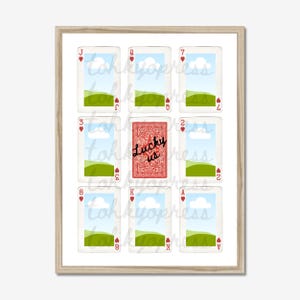 May include: A framed print featuring playing cards with a landscape design. The central card reads "Lucky us" in script. The other cards display hearts and numbers. The artwork is set against a white background.