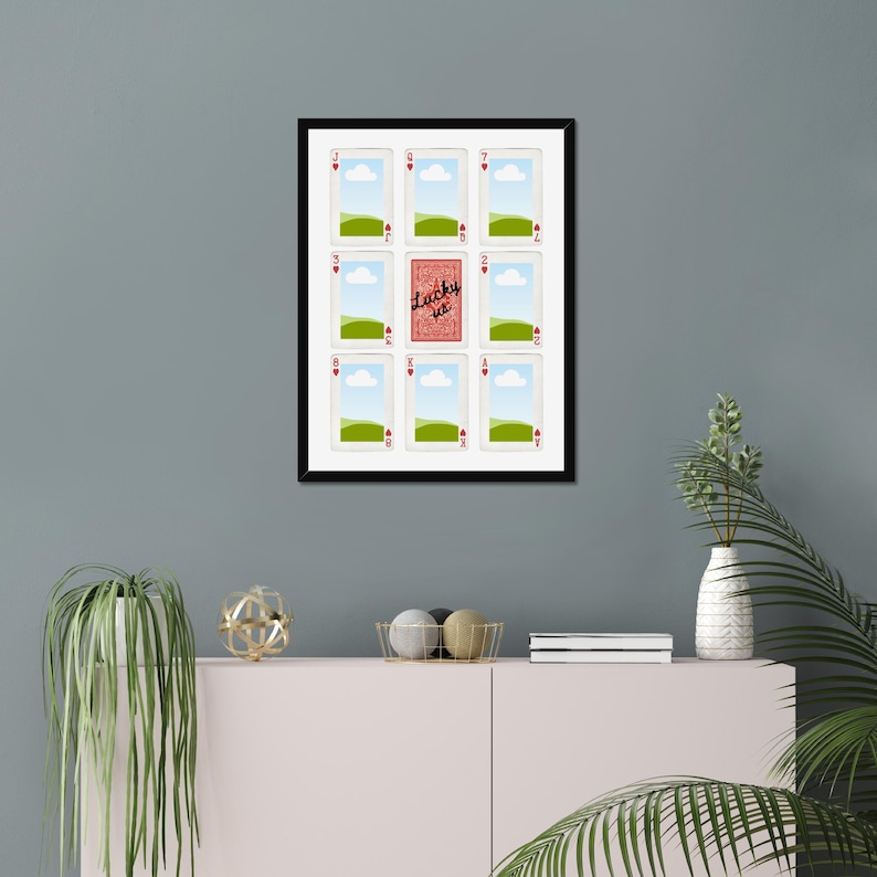 May include: Framed artwork featuring playing cards. The central card is red and reads "Lucky us." The surrounding cards are white with heart symbols and landscape scenes. The frame is black.