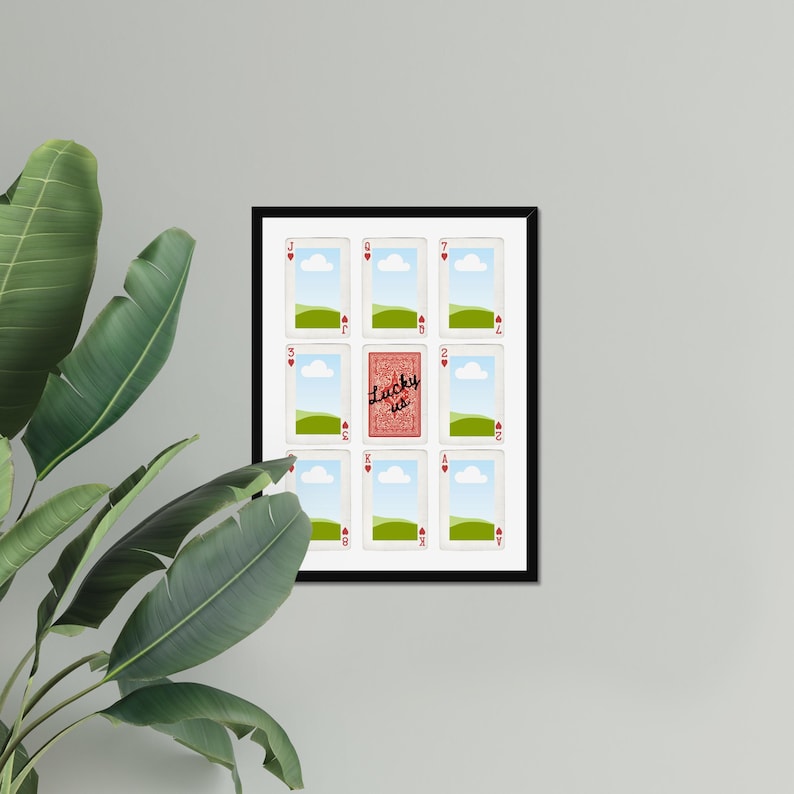 May include: A framed artwork featuring playing cards. The central card is red and reads "Lucky us." The other cards are white with red hearts, green fields, and blue skies. The frame is black.