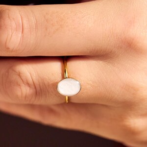 May include: A gold ring with a white oval stone. The ring is on a person's hand, with a blurred background. The ring's band is thin and delicate, and the stone is set in a simple, elegant style. Jewelry.