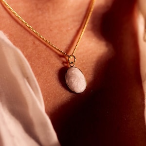 May include: A gold-colored chain necklace with a small, oval, white pendant. The necklace is worn against a white shirt. The pendant has a textured surface and is the focal point of the image.