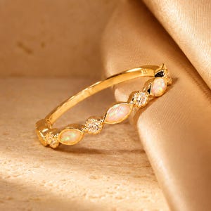 May include: A gold ring with opal and diamond detailing. The ring has a slender band adorned with alternating oval opal stones and small diamond clusters. It is displayed on a textured, light-toned surface and a draped, golden fabric.