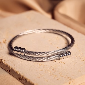 Op de afbeelding: Een zilverkleurige, gedraaide kabelarmband met een wikkelontwerp. De armband ligt op een getextureerd, lichtgekleurd rechthoekig oppervlak, met een zachte, beige stof op de achtergrond. De armband heeft kleine, ronde decoratieve accenten.