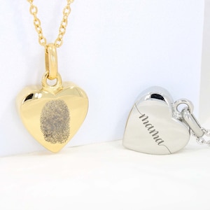Peut inclure: Deux pendentifs en forme de cœur sur des chaînes. L'un est doré avec une empreinte digitale, l'autre est argenté avec le mot "mama" gravé. Les deux pendentifs sont présentés sur un fond blanc.