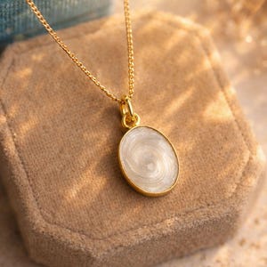 Può includere: Una collana color oro con un pendente ovale. Il pendente presenta un design bianco a spirale e un bordo dorato. La collana è esposta su un portagioie in velluto beige.