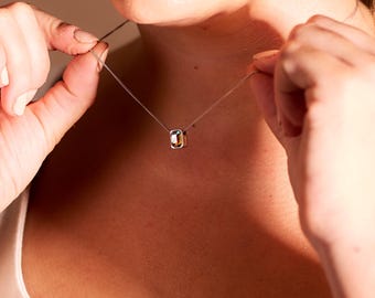 Puede incluir: Un delicado collar de plata con un colgante rectangular con detalles dorados y plateados. Una persona sostiene el collar, mostrando el diseño de la joya y cómo se coloca en el cuello. El fondo es de un tono cálido y neutro.