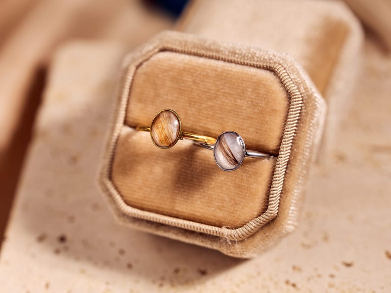 May include: Two rings with oval-shaped, clear resin settings containing hair. One ring has a gold band, the other a silver band. The rings rest in a beige velvet ring box, with a soft-focus background.