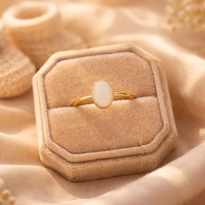 May include: A gold ring with an oval, white gemstone is displayed in a beige velvet ring box. The ring box is on a light peach fabric, with a pair of baby booties in the background. The ring is a piece of jewelry.