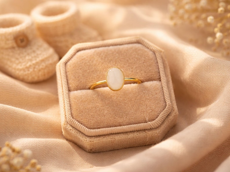 May include: A gold ring with an oval, white gemstone is displayed in a beige velvet ring box. The ring box is on a light peach fabric, with a pair of baby booties in the background. The ring is a piece of jewelry.