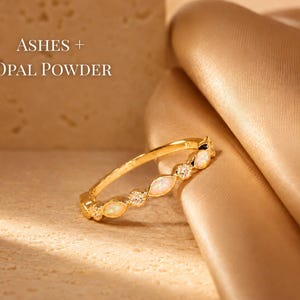 May include: A gold ring with opal and diamond accents. The ring is set against a textured beige surface and a draped, golden fabric. Text in the image reads "ASHES + OPAL POWDER".