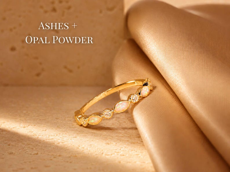 May include: A gold ring with opal and diamond accents. The ring is set against a textured beige surface and a draped, golden fabric. Text in the image reads "ASHES + OPAL POWDER".