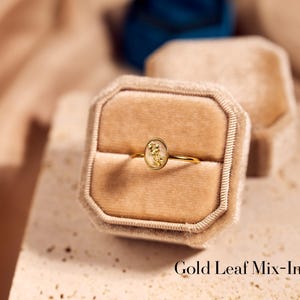 May include: A gold ring with an oval-shaped centerpiece containing gold leaf details, presented in a beige velvet ring box. The ring has a delicate band and is set against a neutral background. The text "Gold Leaf Mix-In" is visible.